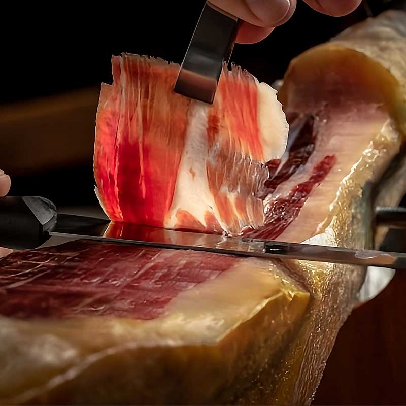 Primer plano del corte manual de una loncha de jamón, mostrando la textura y el brillo de la grasa.